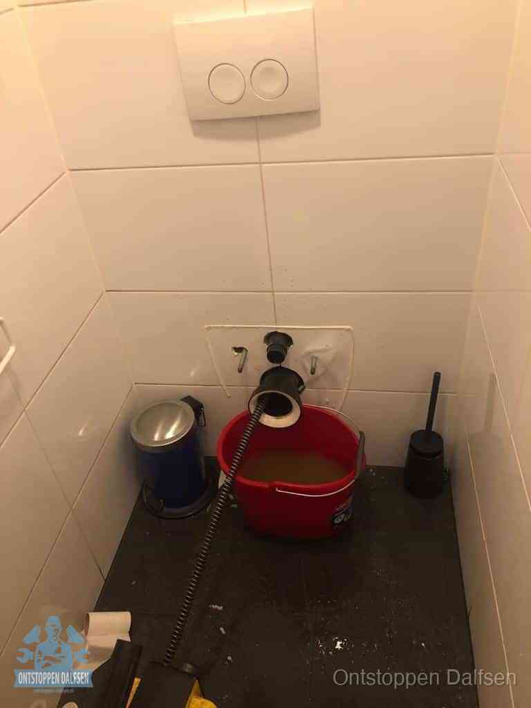 Een ontstopte wand-wc met verwijderde toiletpot waar een ontstoppingsveer in de wanduitgang verdwijnt, het achterhalen waar de verstopping zit in Dalfsen
