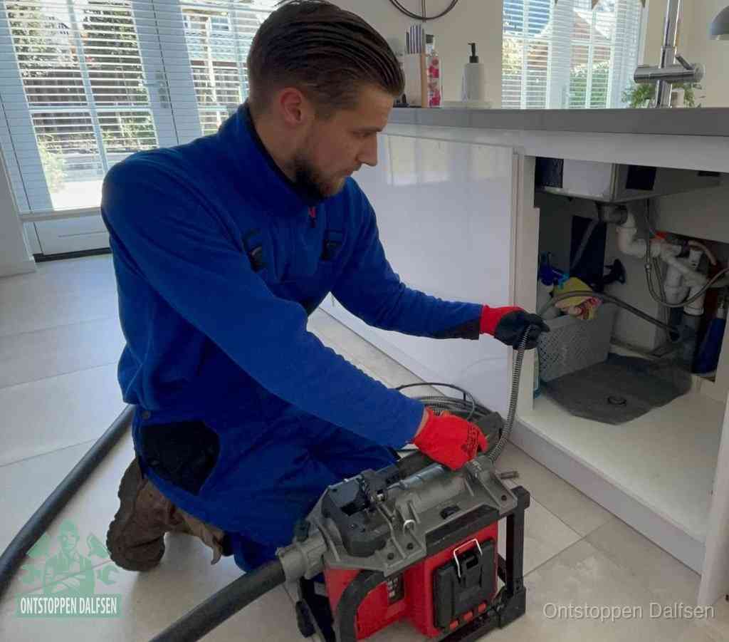 Een monteur met rode handschoenen knielt in een keuken en voert een elektrische ontstoppingsveer in tijdens het professioneel afvoer ontstoppen in Dalfsen