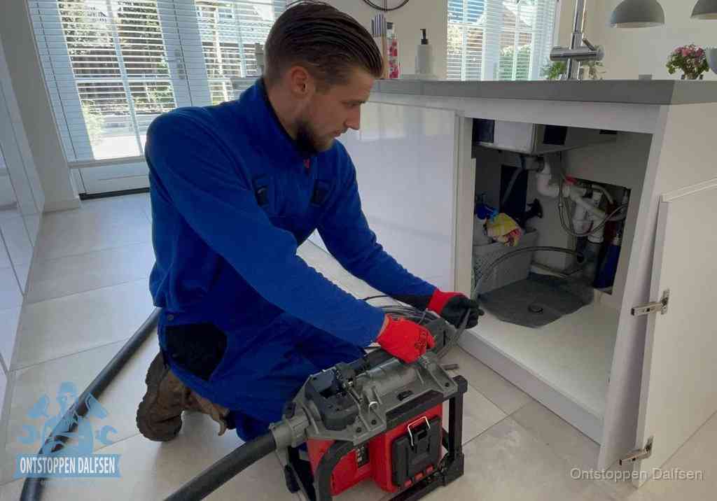 Een monteur voert een professionele elektrische ontstoppingsmachine in de afvoer onder de spoelbak tijdens het gootsteen ontstoppen in Dalfsen